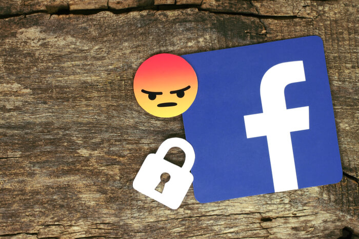Facebook-Logo mit Hängeschloss und wütendem Smiley auf einem hölzernen Hintergrund.