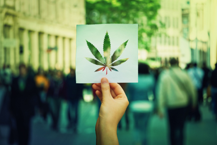 Hand, die ein Papierblatt mit einem Symbol für Marihuana-Blätter auf einem überfüllten Straßenhintergrund hält.